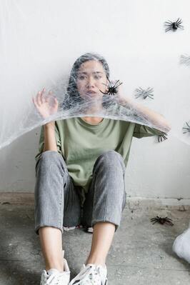 A picture of a scared young woman, wearing a green shirt and blue jeans, trapped behind cobwebs that have spiders on it. She is sitting behind the cobwebs in front of a white wall.